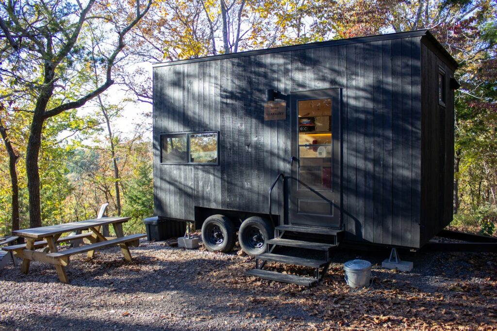 A small dark-sided tiny house with a wooden step and a picnic table in a wooded yard